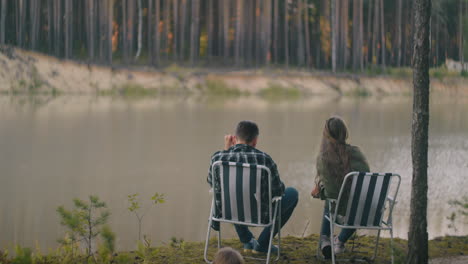 couple-of-hikers-is-admiring-landscape-with-lake-in-forest-man-and-woman-are-sitting-in-chair-in-woodland-camp-and-relaxing